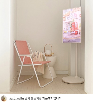 LG 스탠바이미가 코너에 설치된 모습으로, TV 화면에는 거리 풍경 그림이 표시되어 있음. 옆에는 핑크색 접이식 의자와 테이블 위에 담요와 화분이 배치되어 있으며, 하단에는 'yaru_yallu 님의 오늘의집 제품후기입니다.'라는 텍스트가 포함되어 있음.