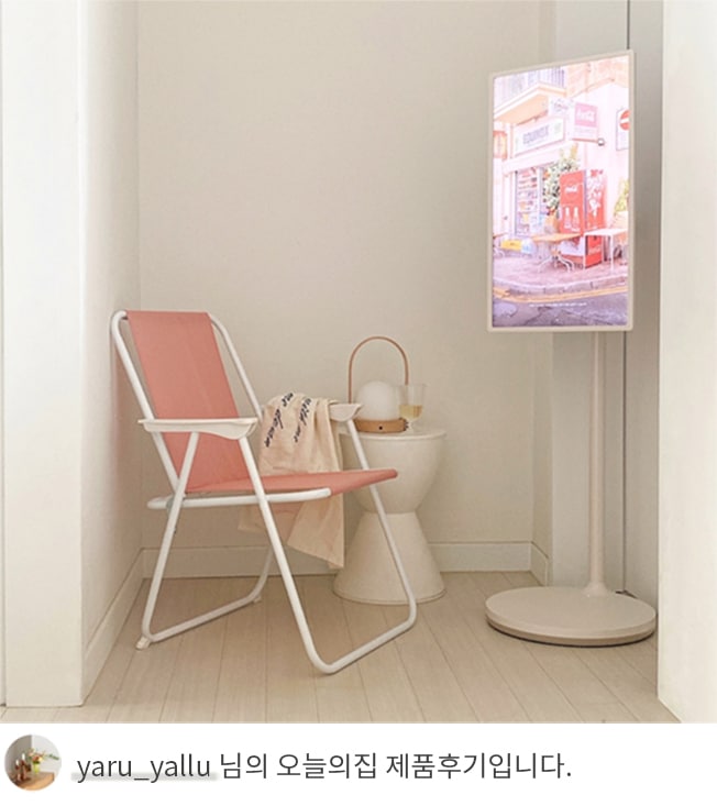 LG 스탠바이미가 코너에 설치된 모습으로, TV 화면에는 거리 풍경 그림이 표시되어 있음. 옆에는 핑크색 접이식 의자와 테이블 위에 담요와 화분이 배치되어 있으며, 하단에는 'yaru_yallu 님의 오늘의집 제품후기입니다.'라는 텍스트가 포함되어 있음. 
