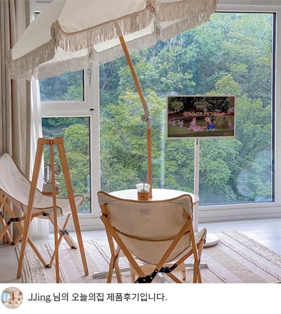 큰 창문 앞에 놓인 LG 스탠바이미와 야외용 의자, 테이블이 함께 배치된 모습입니다. 테이블 위에는 음료가 놓여 있으며, 창문 너머로는 푸르른 숲의 풍경이 펼쳐져 있습니다. 하단에는 'JJing 님의 오늘의집 제품후기입니다.'라는 텍스트가 포함되어 있음.