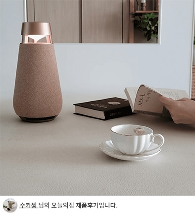 테이블 위에 놓인 커피잔과 받침, 책 두 권, 그리고 무드등 옆에서 책을 읽는 손의 모습. 배경에는 선반과 식물이 보이며, 하단에 '수카짱 님의 오늘의집 제품후기입니다.'라는 텍스트가 포함되어 있음.
