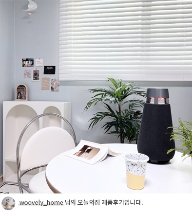 LG 엑스붐 360 스피커가 흰색 둥근 테이블 위에 놓여 있는 모습. 테이블 옆에는 화분과 의자가 있으며, 창문에는 블라인드가 설치되어 있음. 하단 텍스트는 'woovely_home 님의 오늘의집 제품후기입니다.' 로 구성됨.