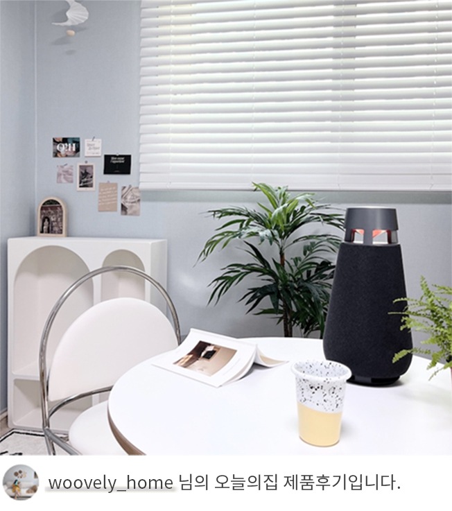 LG 엑스붐 360 스피커가 흰색 둥근 테이블 위에 놓여 있는 모습. 테이블 옆에는 화분과 의자가 있으며, 창문에는 블라인드가 설치되어 있음. 하단 텍스트는 'woovely_home 님의 오늘의집 제품후기입니다.' 로 구성됨.