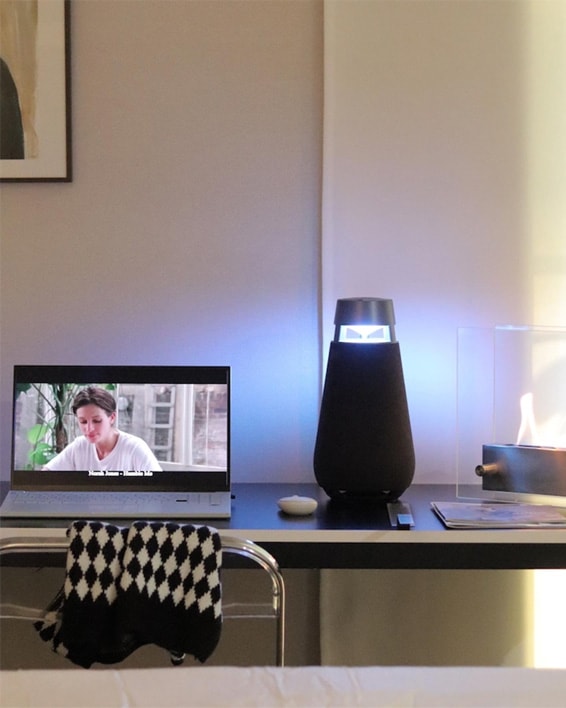 LG 엑스붐 360 스피커가 책상 위에 놓여 있으며, 노트북 화면에는 영화를 재생 중인 모습이 보입니다. 주변에는 현대적인 인테리어 소품과 함께 따뜻한 분위기를 연출하고 있습니다.