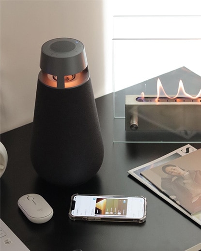 LG 엑스붐 360 스피커가 작은 테이블 위에 놓여 있고, 스마트폰과 함께 아늑한 실내 공간에 배치된 모습입니다. 주변에는 미니 화로와 소품이 어우러져 따뜻하고 감성적인 분위기를 자아냅니다.