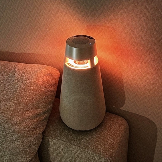 LG 엑스붐 360 스피커가 따뜻한 조명을 켠 채 소파 옆에 배치된 모습.