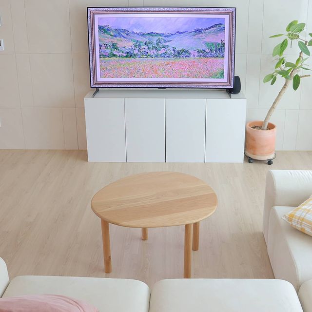 밝은 거실에 흰색 소파와 원형 우드 테이블이 배치되어 있음. TV가 거실 벽에 설치되어 있고, 인상파 풍경화가 화면에 표시됨. 옆에는 화분에 심어진 키 큰 식물이 있음.