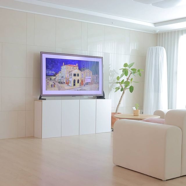 밝은 거실에 흰색 소파와 우드 테이블이 배치되어 있음. TV 화면에는 야경이 담긴 인상파 그림이 나옴. 옆에는 화분에 심어진 키 큰 식물이 있음.