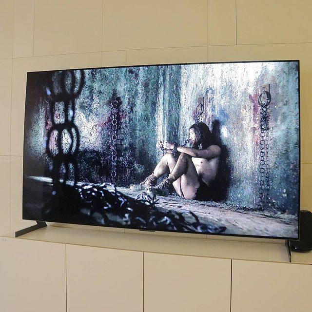 거실 벽에 설치된 TV 화면에 사슬에 묶인 남성이 어두운 감옥 같은 공간에 앉아 있는 장면이 나옴. TV는 낮은 흰색 수납장 위에 놓여 있음.