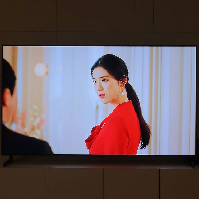 거실 벽에 설치된 TV가 화면에 붉은색 의상을 입은 여성이 뒤를 돌아보고, 왼쪽에는 검은색 정장을 입은 남성의 뒷모습이 나옴.