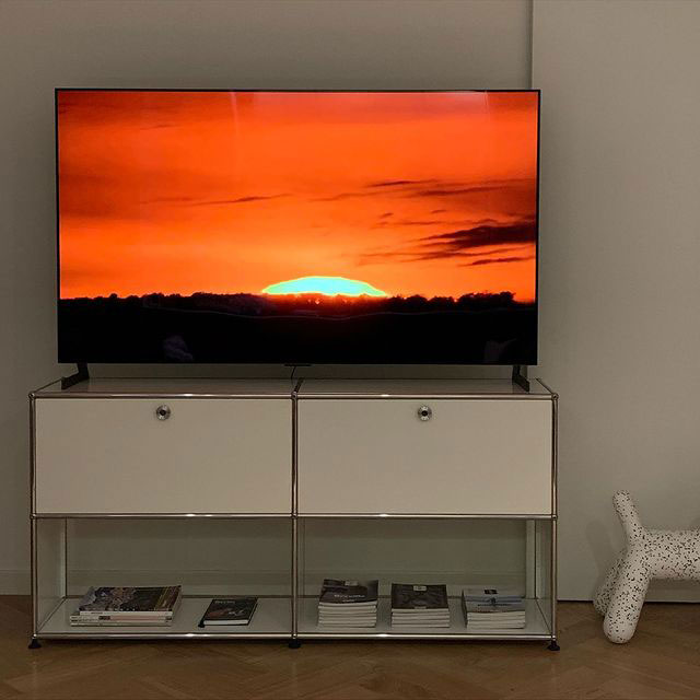 TV 화면에 붉은 노을이 지는 풍경이 보이며, TV는 화이트 수납장 위에 놓여 있음. 아래에는 책과 잡지가 정리되어 있으며, 옆에는 강아지 모양의 장식품이 있음. 배경에는 심플한 디자인의 벽과 나무 바닥이 있음.