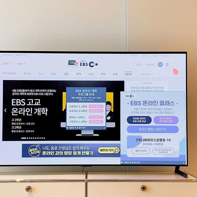 TV 화면에 EBS 온라인 클래스와 관련된 웹페이지가 표시되어 있음. 화면에는 EBS 고교 온라인 개학과 EBS 온라인 클래스 안내 문구가 있으며, 온라인 강의 일정과 무료 이용 안내가 포함됨. TV는 화이트 수납장 위에 놓여 있음.
