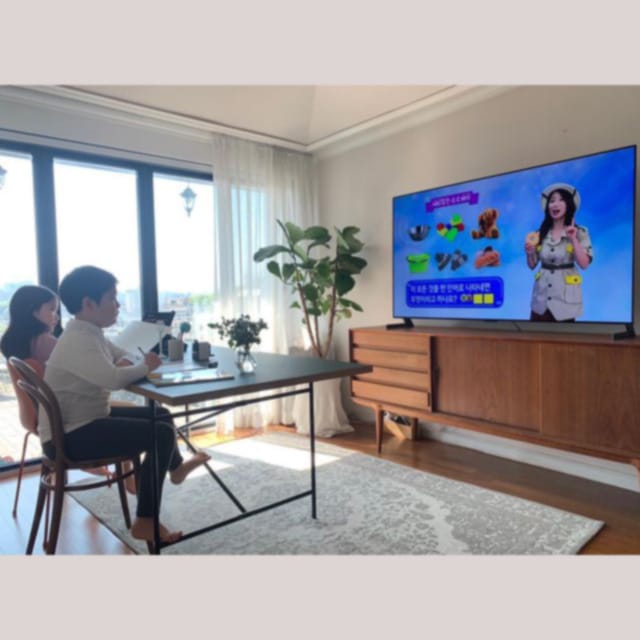 거실에서 어린이가 책상에 앉아 TV로 온라인 수업을 시청하며 공부하는 모습. TV 화면에는 교육 프로그램이 재생 중이며, 배경에는 큰 창과 화분, 우드 톤 가구가 있음.