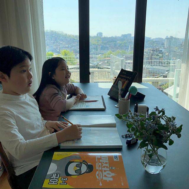 창가 책상에서 어린이 두 명이 교재를 펼쳐두고 학습하는 모습. 창밖으로 도심 전경이 보이며, 책상 위에는 학습 도구와 화분이 놓여 있음.