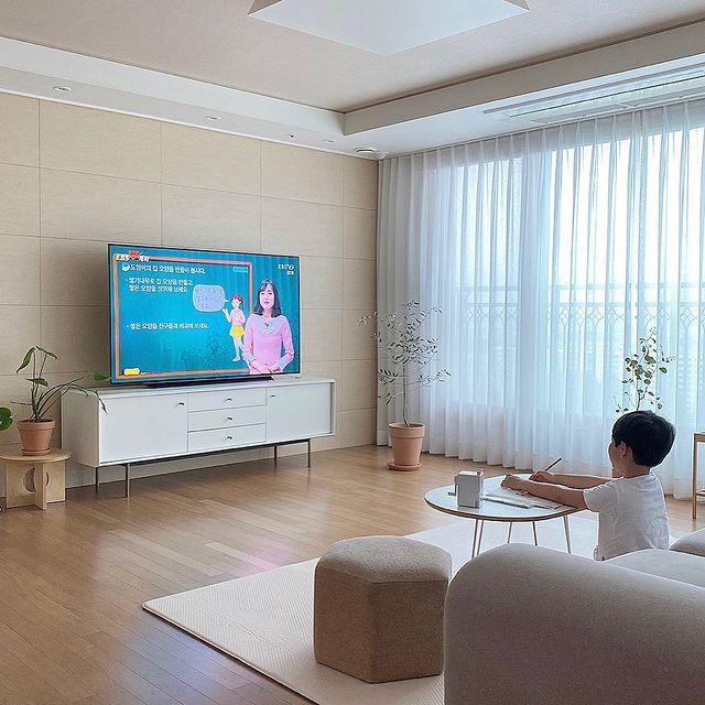 거실에 놓인 대형 TV에서 온라인 수업이 재생되고 있으며, 아이가 원형 테이블에 앉아 필기를 하고 있음. 배경에는 밝은 자연광이 들어오는 창가에는 얇은 커튼과 화분이 배치되어 있음.