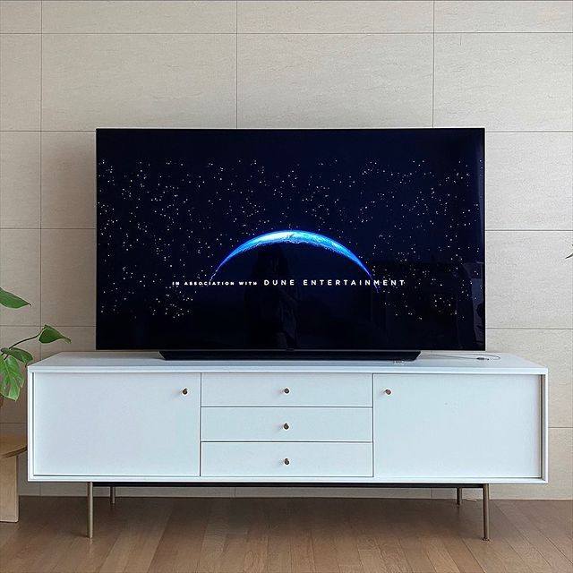 거실의 흰색 거실장 위에 배치된 TV에서 DUNE ENTERTAINMENT 로고 화면이 표시됨. 배경에는 베이지톤 벽과 작은 화분이 있어 정돈된 분위기임.