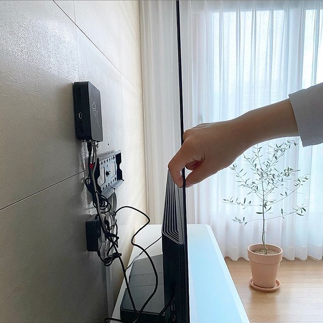 초슬림 디자인의 TV를 손으로 살짝 들어 올리는 모습. 벽에 부착된 케이블 정리함과 연결된 선들이 보이며, 배경에는 커튼이 드리워진 창과 화분이 놓인 밝은 거실이 있음.