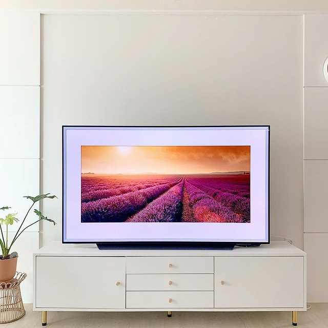 흰색 거실장 위에 놓인 TV 화면에 연보라색 라벤더 밭이 펼쳐진 풍경이 표시됨. 배경에는 깔끔한 흰 벽면과 왼쪽에 작은 화분이 있음.
