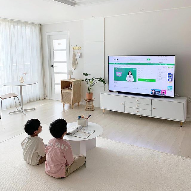 흰색 거실에서 두 아이가 작은 원형 테이블 앞에 앉아 TV로 EBS 온라인 학습을 시청하며 공부하고 있음. 배경에는 큰 창과 흰 커튼, 작은 테이블과 의자가 있음.