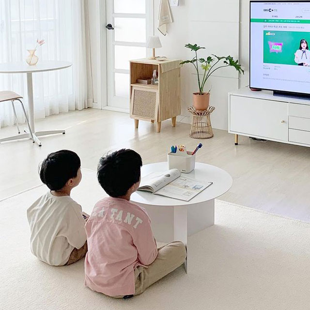 흰색 거실에서 두 아이가 작은 원형 테이블 앞에 앉아 TV로 EBS 온라인 학습을 시청하며 공부하고 있는 모습 클로즈업. 배경에는 큰 창과 흰 커튼, 작은 테이블과 의자가 있음.
