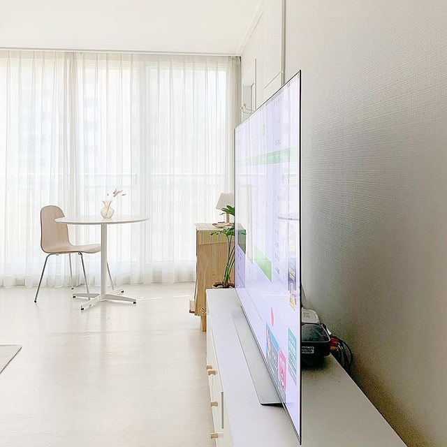 옆면에서 본 초슬림 TV가 거실장 위에 배치됨. 배경에는 커튼이 있는 큰 창과 원형 테이블, 의자가 있음.