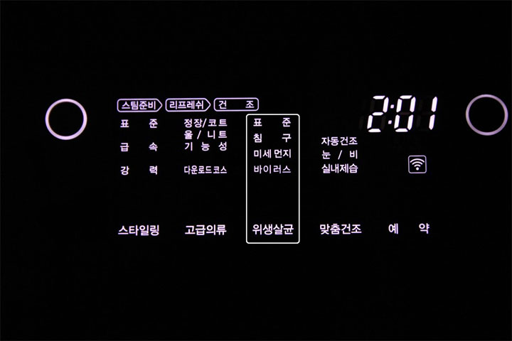 어두운 배경에서 LG 스타일러의 디지털 패널이 밝게 빛나는 모습으로, '위생살균'과 '바이러스코스' 등 다양한 기능과 설정 시간이 또렷하게 표시되어 있음