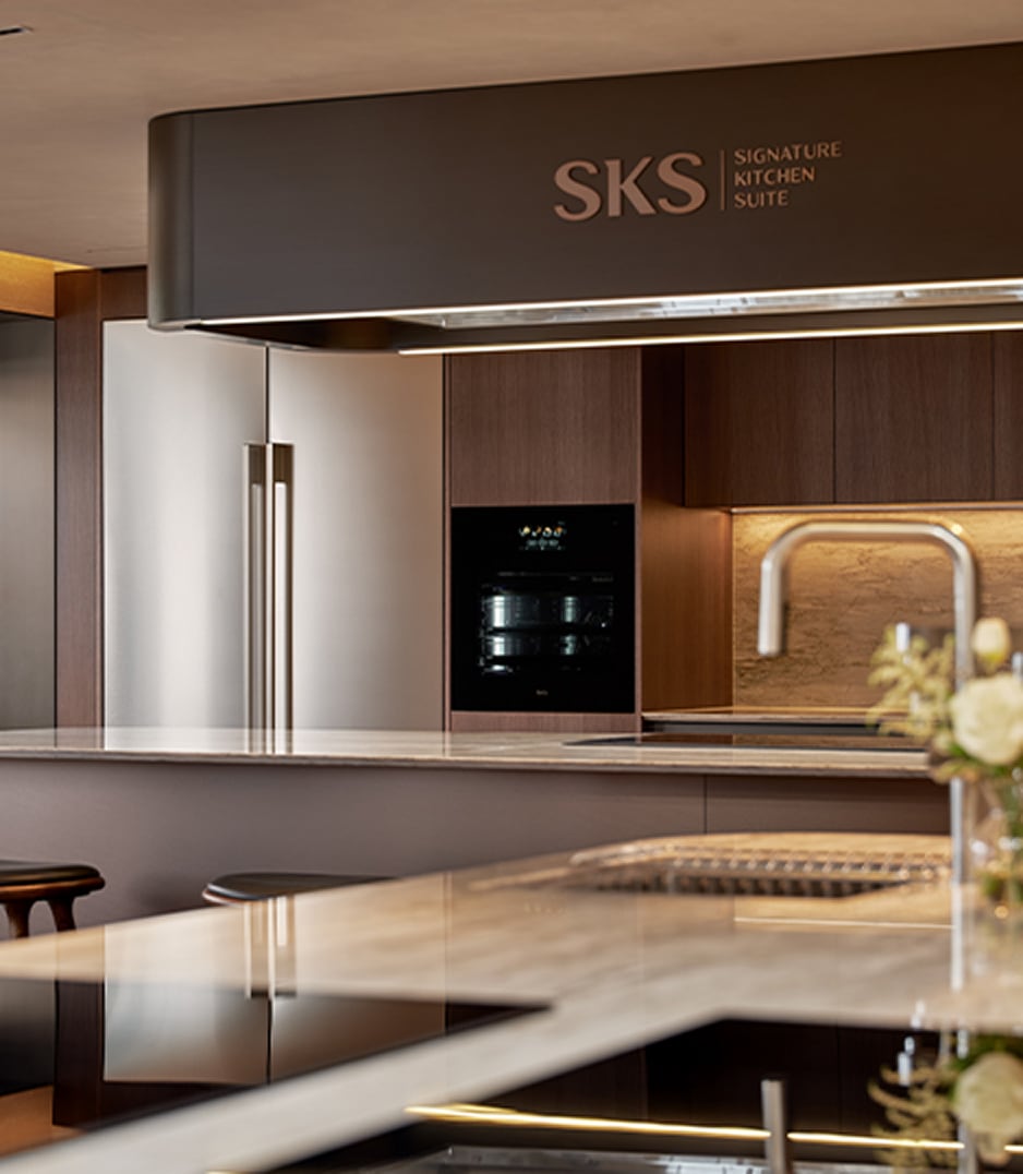 SKS(Signature Kitchen Suite) 로고가 보이는 고급 주방 공간. 스테인리스 양문형 냉장고와 오븐이 나란히 설치되어 있으며, 마블 소재의 조리대와 수전, 우드 패널 벽면이 조화를 이루고 있음.