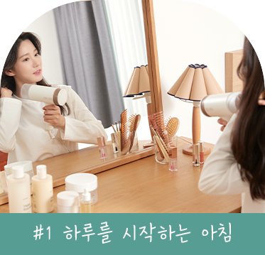 화장대 앞에서 헤어드라이어를 사용하며 하루를 시작하는 여성의 모습. 화장대 위에는 스킨케어 제품과 빗이 정리되어 있고, 배경에는 따뜻한 조명이 더해져 있다. 이미지 하단에는 '#1 하루를 시작하는 아침'이라는 텍스트가 포함되어 있다.