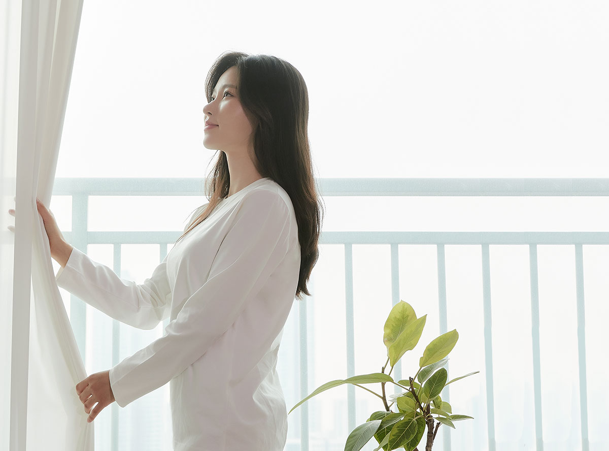 창문을 바라보며 커튼을 걷고 있는 여성이 밝은 아침 햇살을 느끼는 모습. 창가 옆에는 푸른 잎의 화분이 놓여 있음.