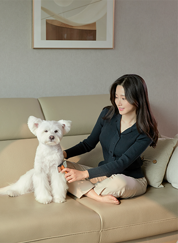 소파에 앉아 흰색 강아지의 털을 강아지 빗으로 빗어주고 있는 여성. 강아지가 편안한 표정으로 정면을 바라보고 있는 모습.