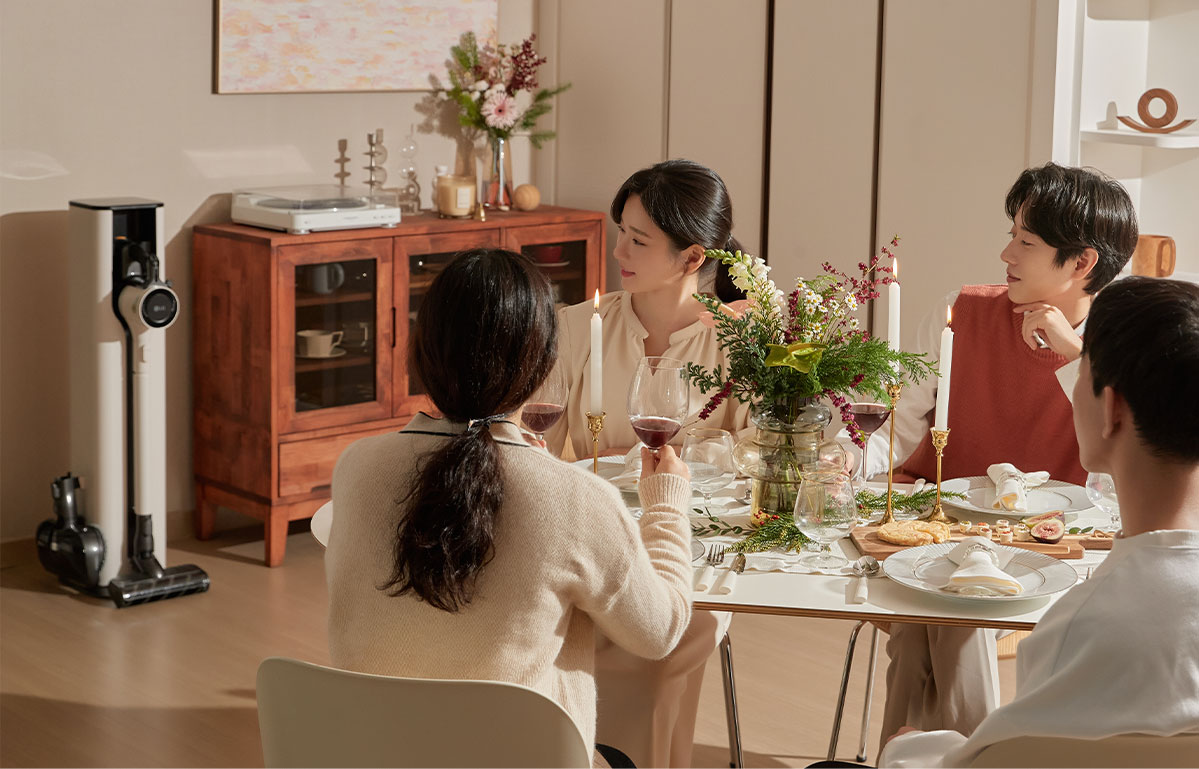 원목 찬장 옆에 놓인 LG 코드제로 A9S 오브제컬렉션 청소기를 배경으로, 꽃으로 꾸며진 식탁에 앉아 와인을 마시며 대화를 나누는 네 명의 사람들.