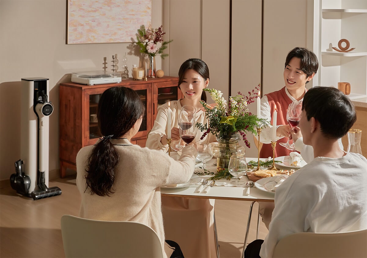 식탁에 앉아 와인을 들고 건배하며 대화를 나누는 네 명의 사람들. 꽃과 촛불로 장식된 식탁이 따뜻하고 아늑한 분위기를 연출.