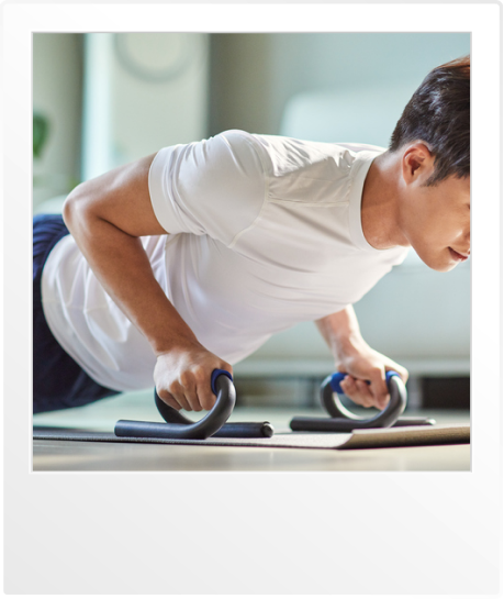 푸쉬업 바를 이용해 집에서 팔굽혀펴기 운동을 하는 남성. 흰색 반팔 티셔츠와 운동복 바지를 착용하고 있으며, 바닥에는 요가 매트가 깔려 있습니다. 밝은 실내 환경에서 운동에 집중하는 모습입니다.