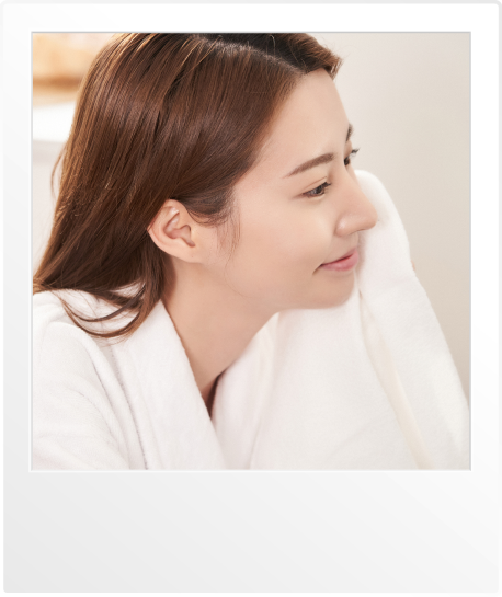 흰색 목욕가운을 입은 여성이 부드러운 타월을 얼굴에 대고 미소 짓고 있는 모습. 깨끗하고 보송보송한 세탁물의 촉감을 즐기는 장면을 보여줍니다.