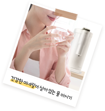 핑크색 셔츠를 입은 여성이 투명한 유리컵에 담긴 물을 두 손으로 들고 있는 모습으로, 배경에는 정수기가 놓여 있으며 '건강한 미네랄이 살아 있는 물 마시기'라는 문구가 있는 이미지