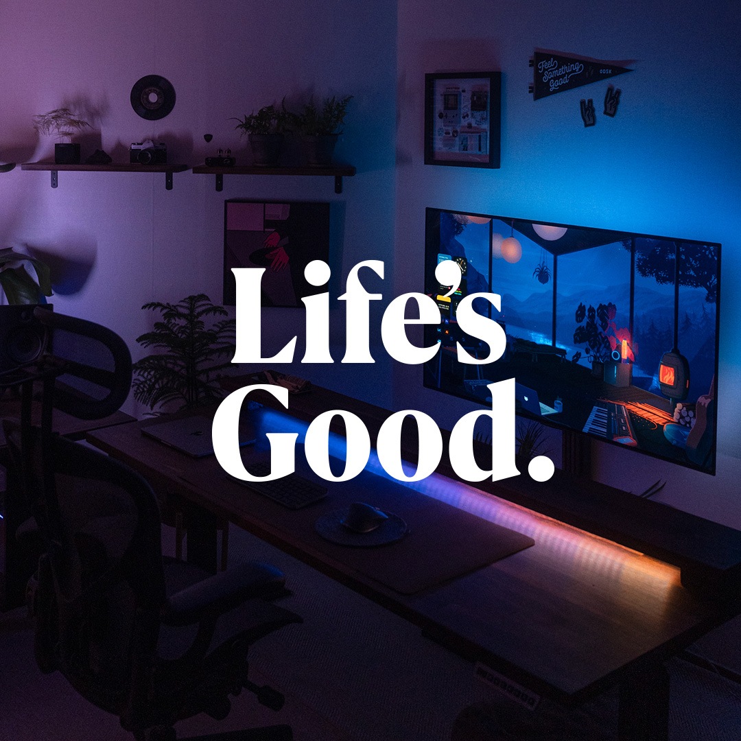 LG OLED TV를 활용한 감성적인 개인 공간으로, 어두운 조명 속에서 블루와 오렌지 네온 라이트가 따뜻한 분위기를 연출하는 모습. 대형 화면에는 창가 너머 숲이 보이는 아늑한 작업실 배경의 애니메이션 장면이 표시되어 있으며, 원목 책상 위에는 키보드, 마우스, 컵 등이 정리되어 차분한 공간감을 형성. 벽에는 선반과 소품, 식물이 배치되어 있으며, 화면 중앙에는 'Life’s Good.' 문구가 강조된 이미지.