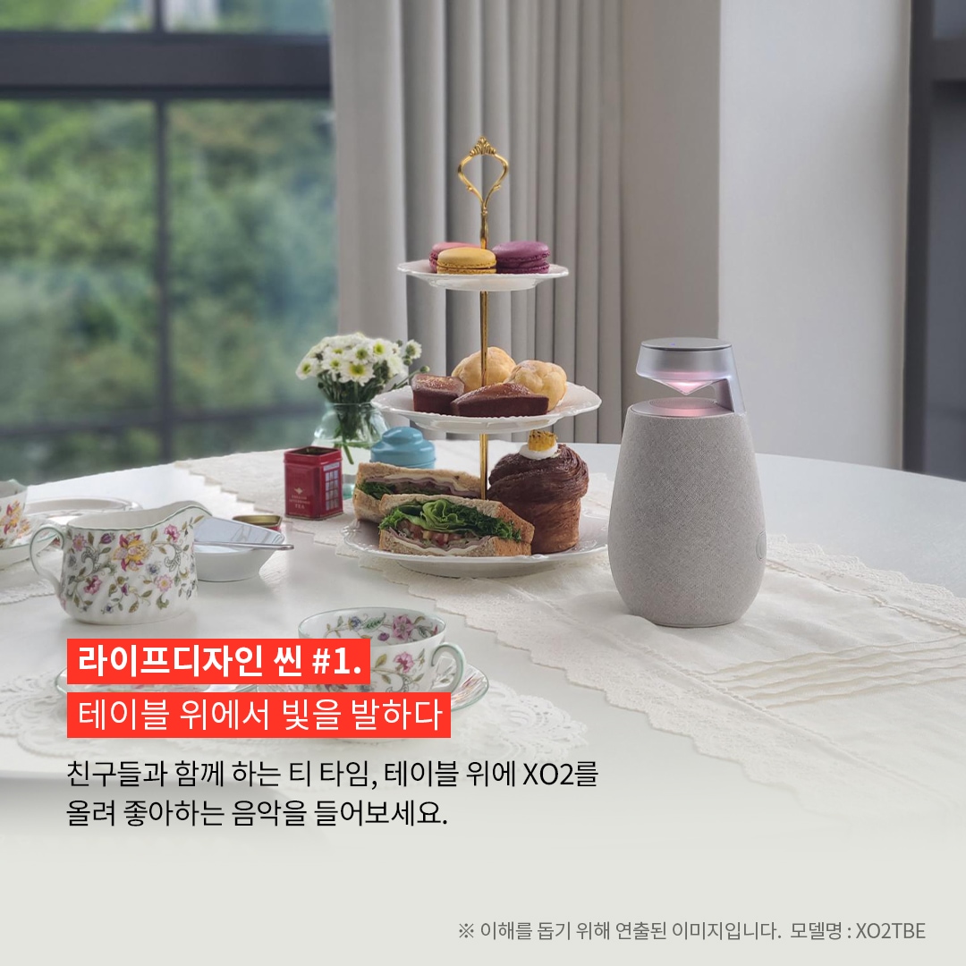 라이프디자인 씬 1.테이블 위에서 빛을 발하다 친구들과 함께하는 티 타임, 테이블 위에 XO2를 올려 좋아하는 음악을 들어보세요. 공간의 분위기가 한결 부드러워질 거예요. ※ 이해를 돕기 위해 연출된 이미지입니다. 모델명: XO2TBE 문구 표시됨. 배경은 레이스 테이블보 위에 차와 다양한 디저트가 담긴 3단 접시가 놓인 흰색 원형 테이블. 오른쪽에는 붉은빛으로 점등된 엑스붐 360 스피커가 있고, 배경으로는 창문 너머로 푸른 자연이 보임.