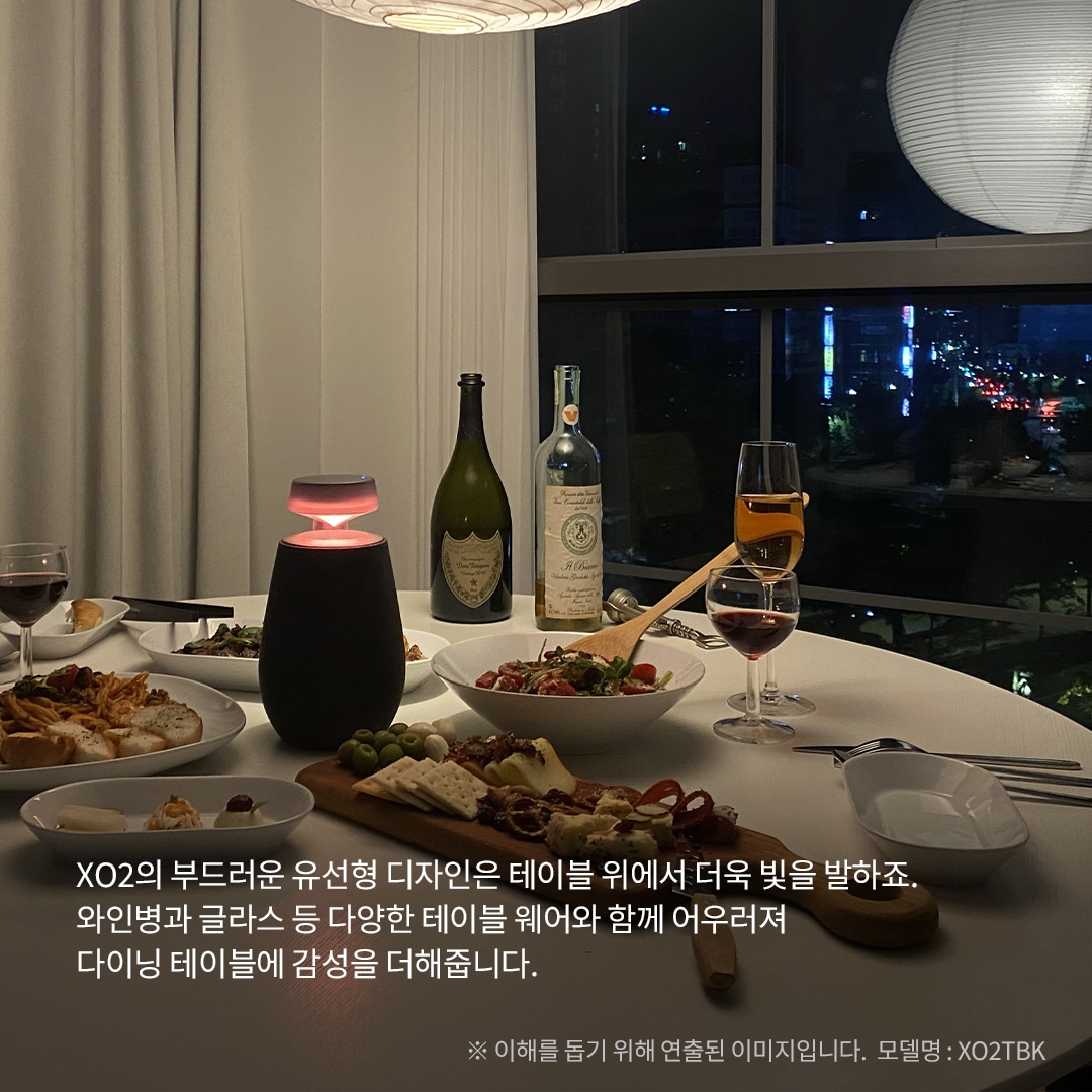 XO2의 부드러운 유선형 디자인은 테이블 위에서 더욱 빛을 발하죠. 와인병과 글라스 등 다양한 테이블웨어와 함께 어우러져 다이닝 테이블에 감성을 더해줍니다. ※ 이해를 돕기 위해 연출된 이미지입니다. 모델명 : XO2TBK 문구 표시됨. 배경은 야경이 보이는 창문 앞에 흰색 테이블이 놓여 있으며, 테이블 위에는 붉은빛으로 빛나는 엑스붐 360 스피커와 다양한 음식이 준비된 접시들, 와인 두 병과 잔이 있음.