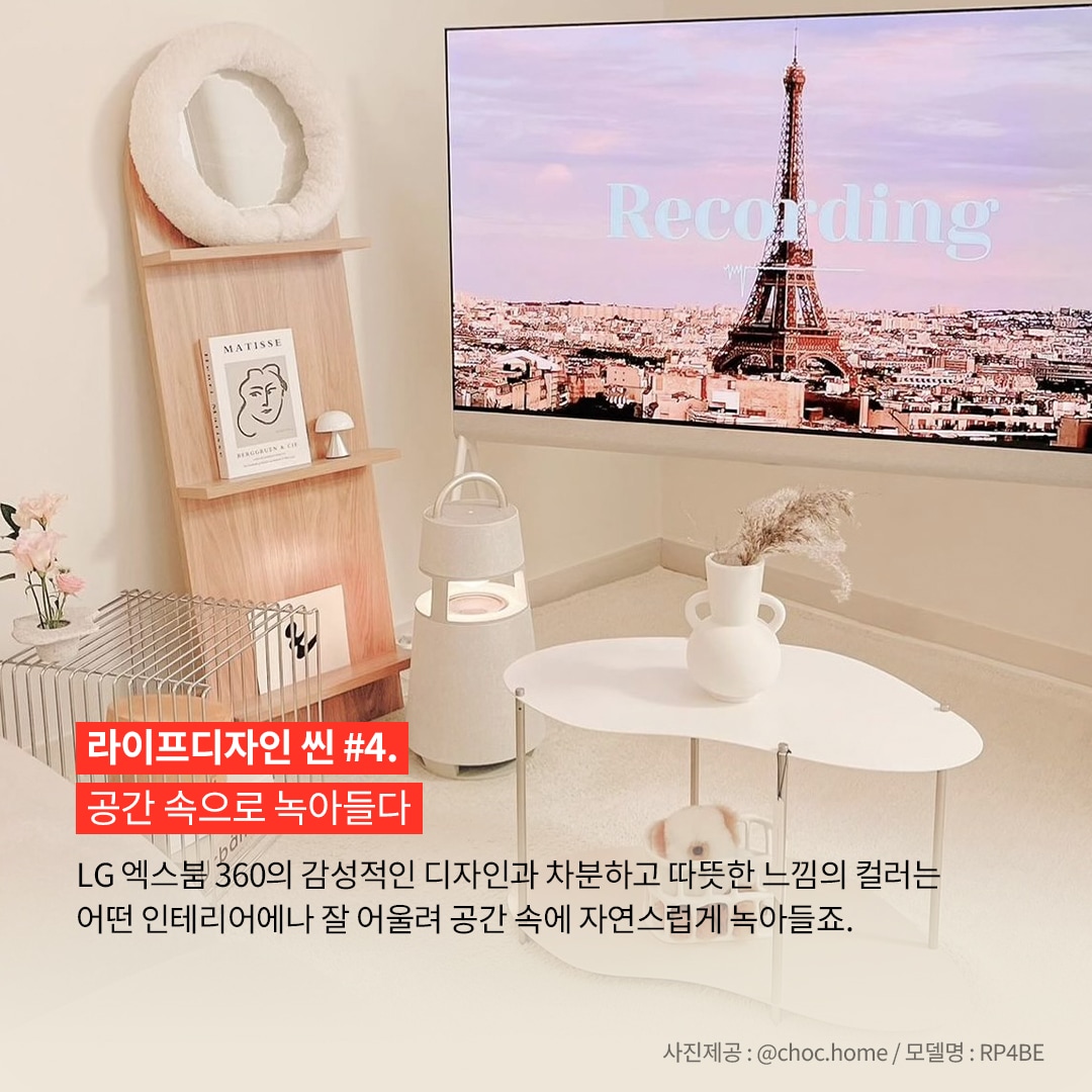 라이프디자인 씬 4. 공간 속으로 녹아들다 LG 엑스붐 360의 감성적인 디자인과 차분하고 따뜻한 느낌의 컬러는 어떤 인테리어나 잘 어울려 공간 속에 자연스럽게 녹아들죠. 사진제공: @choc.home / 모델명: RP4BE 문구 표시됨. 배경은 밝은 실내에서, 흰색 원형 테이블과 나무 선반, 그리고 선반 위의 장식들이 보임. 선반에는 그림과 소품이 있으며, 바닥에는 엑스붐 360 스피커가 놓여 있음. 대형 TV에서 에펠탑이 보이는 화면이 나오고 있음.