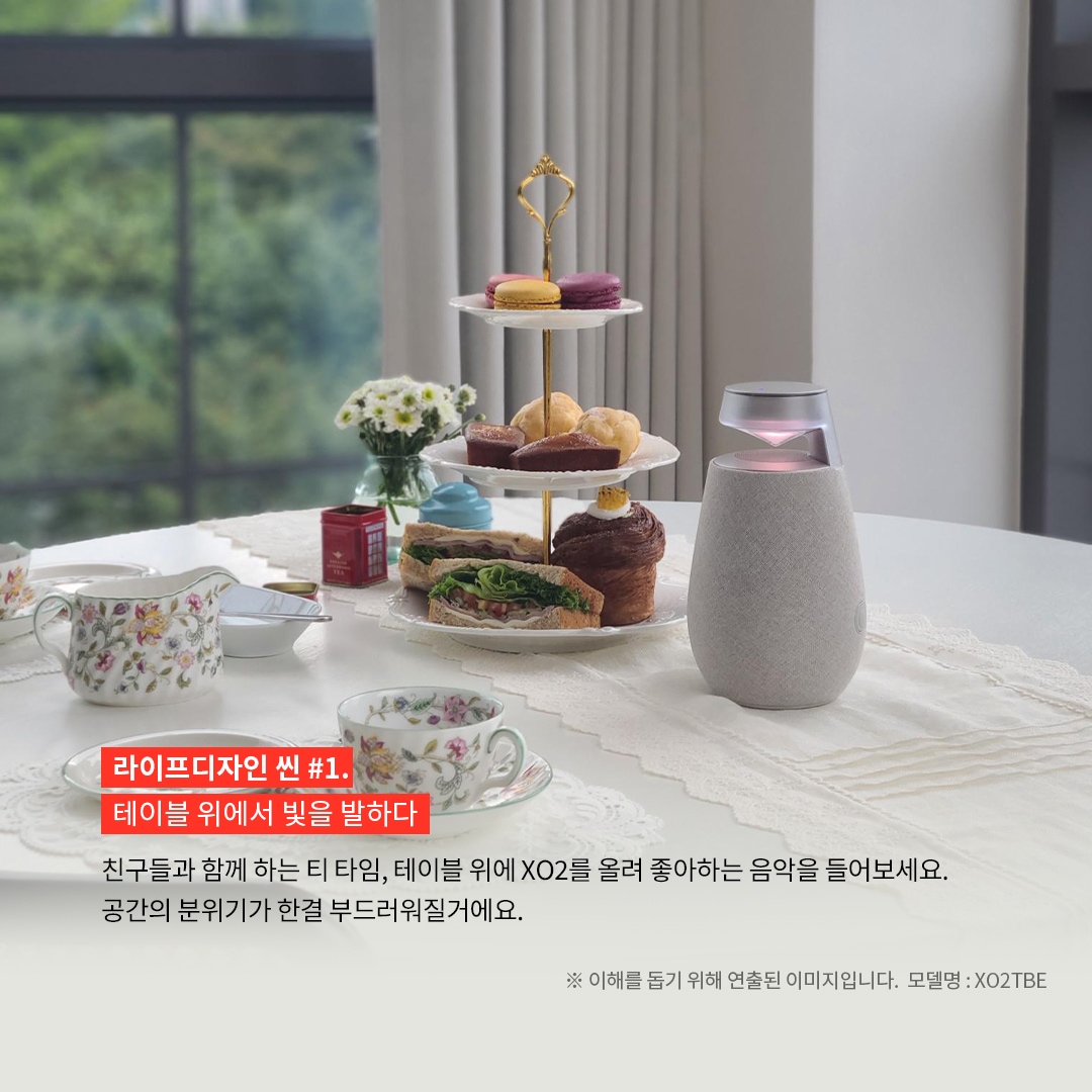 라이프디자인 씬 1.테이블 위에서 빛을 발하다 친구들과 함께하는 티 타임, 테이블 위에 XO2를 올려 좋아하는 음악을 들어보세요. 공간의 분위기가 한결 부드러워질 거예요. ※ 이해를 돕기 위해 연출된 이미지입니다. 모델명: XO2TBE 문구 표시됨. 배경은 레이스 테이블보 위에 차와 다양한 디저트가 담긴 3단 접시가 놓인 흰색 원형 테이블. 오른쪽에는 붉은빛으로 점등된 엑스붐 360 스피커가 있고, 배경으로는 창문 너머로 푸른 자연이 보임.