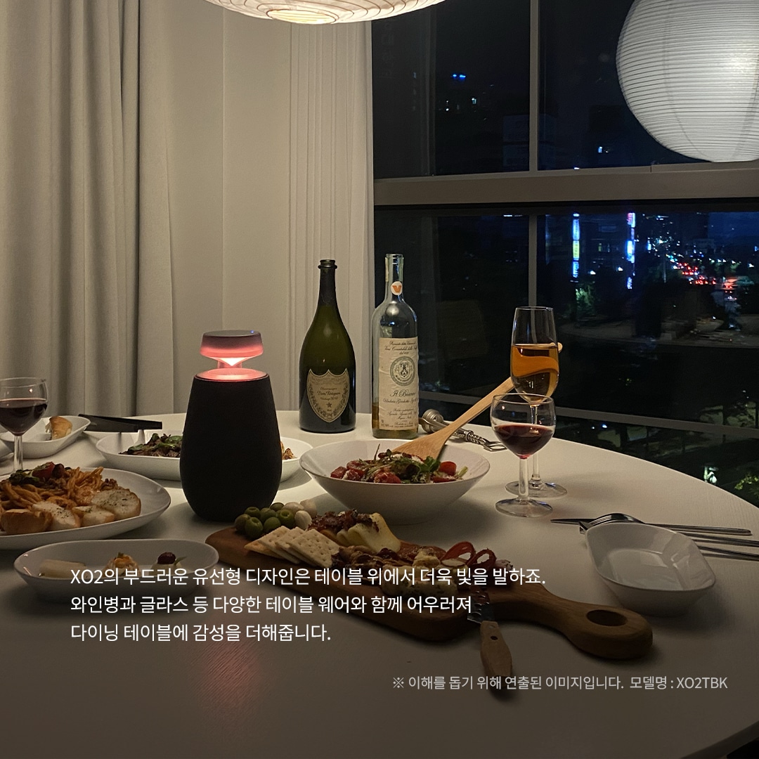 XO2의 부드러운 유선형 디자인은 테이블 위에서 더욱 빛을 발하죠. 와인병과 글라스 등 다양한 테이블웨어와 함께 어우러져 다이닝 테이블에 감성을 더해줍니다. ※ 이해를 돕기 위해 연출된 이미지입니다. 모델명 : XO2TBK 문구 표시됨. 배경은 야경이 보이는 창문 앞에 흰색 테이블이 놓여 있으며, 테이블 위에는 붉은빛으로 빛나는 엑스붐 360 스피커와 다양한 음식이 준비된 접시들, 와인 두 병과 잔이 있음.