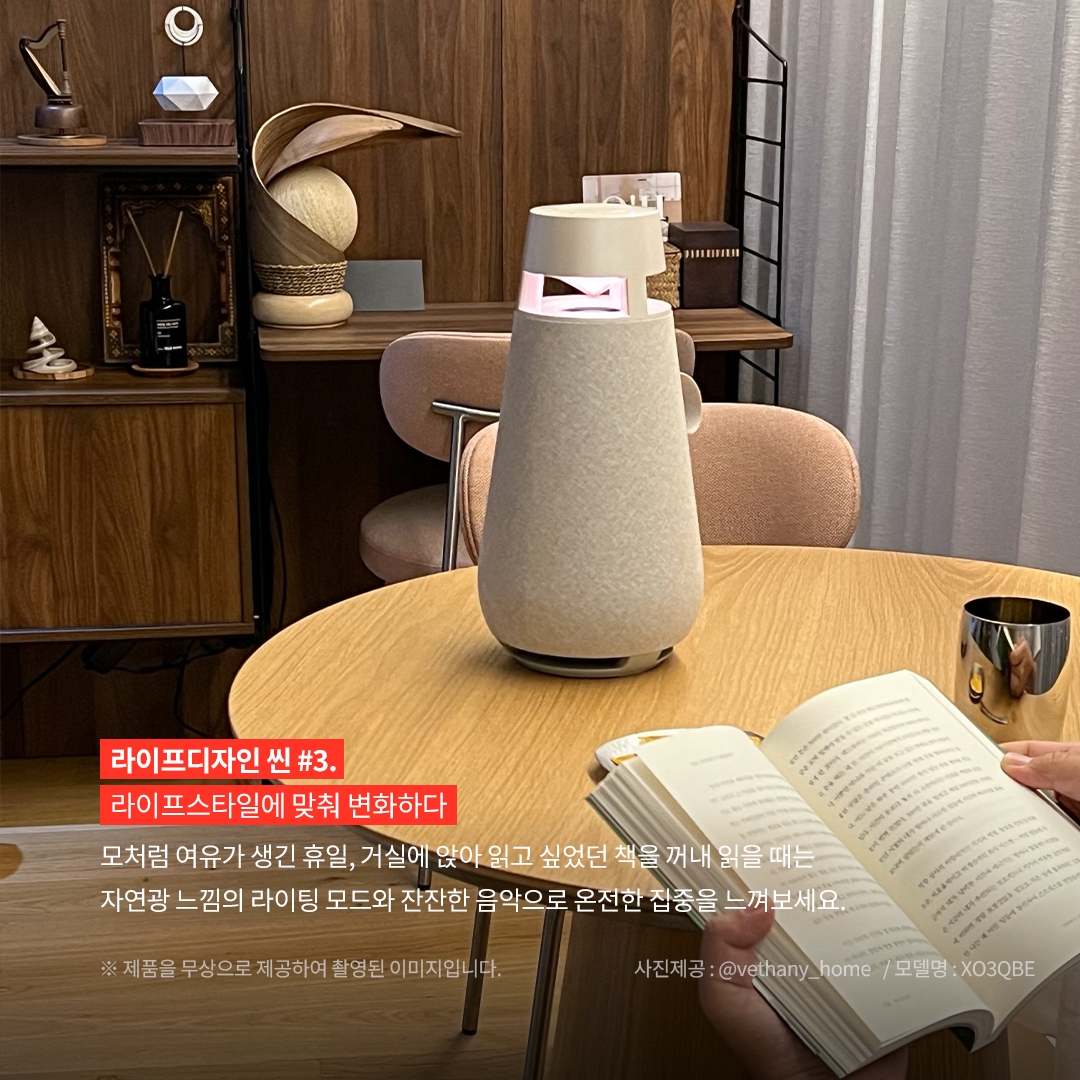 라이프디자인 씬 3. 라이프스타일에 맞춰 변화하다 모처럼 여유가 생긴 휴일, 거실에 앉아 읽고 싶었던 책을 꺼내 읽을 때는 자연광 느낌의 라이팅 모드와 잔잔한 음악으로 온전한 집중을 느껴보세요. ※ 제품을 무상으로 제공하여 촬영된 이미지입니다. 사진제공: @vethany_home / 모델명: XO3QBE 문구 표시됨. 배경은 목재 테이블 위에 모래색 엑스붐 360 스피커가 놓여 있으며, 조명이 은은한 붉은빛을 내고 있음. 앞쪽에는 책을 읽는 손이 보이며, 배경에는 목재 책장과 장식품, 커튼이 있는 아늑한 실내 공간이 보임.