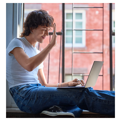 창가에 앉은 인물이 노트북으로 영상 통화 중 손을 흔들며 인사하는 모습. 자연스럽고 편안한 사용자 경험을 표현함.