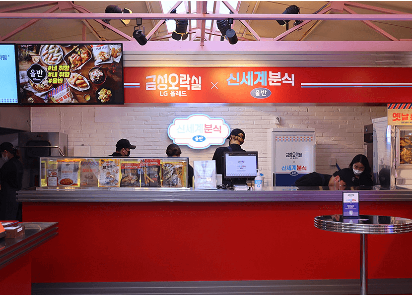붉은색을 메인으로 한 실내 푸드 부스의 모습으로, 상단에는 '금성오락실 LG 올레드 × 신세계분식'이라는 문구와 함께 로고가 있음. 부스 전면에는 다양한 분식 메뉴 사진이 담긴 디스플레이가 놓여 있으며, 부스 안쪽에는 직원들이 업무를 보고 있는 모습