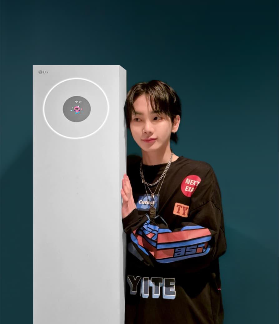 어두운 색상의 캐주얼 의상을 입은 가수 샤이니 키가 스탠드형 에어컨 옆에 기대어 서 있음. 공기청정기의 원형 디스플레이에는 귀여운 캐릭터가 표시됨.