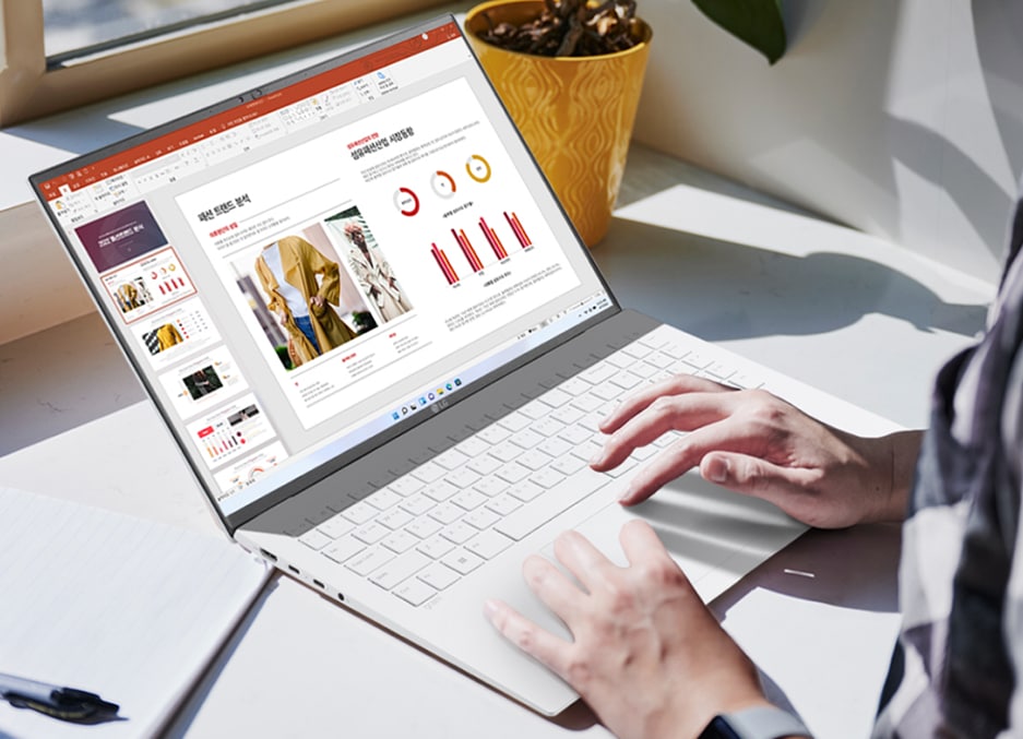 창가에서 한 사람이 화이트 색상의 LG 노트북을 사용하고 있으며, 화면에는 PowerPoint 프레젠테이션이 열려 있습니다. 노트북 옆에는 노트와 펜이 놓여 있으며, 배경에는 노란색 화분이 놓여 있습니다.