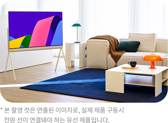 거실 공간에 배치된 세련된 디자인의 TV가 스탠드형 거치대에 놓여 있다. TV 화면에는 추상적인 색상의 그래픽이 표시되어 있으며, 옆에는 크림색 소파와 테이블, 주황색 조명이 놓여 있다.
