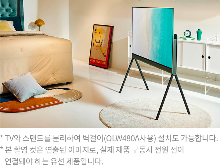 따뜻한 톤의 침실에 스탠드형 TV가 배치된 모습. TV 화면에는 바다가 펼쳐진 장면이 표시되어 있으며, 침대 옆에는 베이지 색상의 러그와 작은 테이블이 있음.