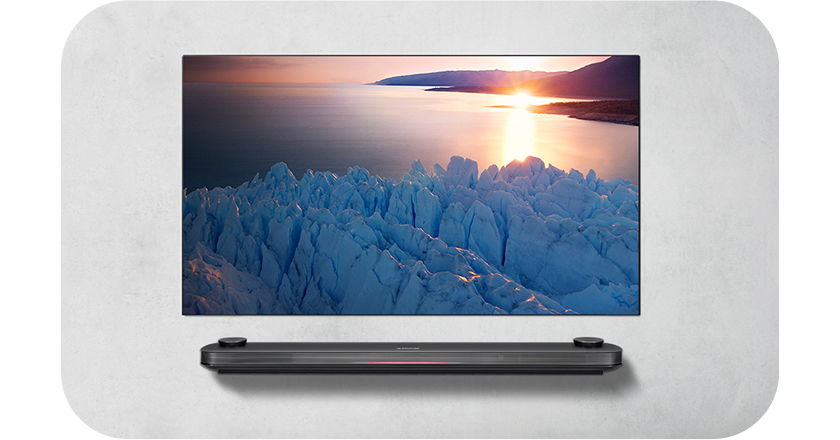 벽에 설치된 LG TV 화면에 빙하와 해안선을 따라 해가 지는 장면이 선명하게 표시됨. TV 아래에는 얇은 사운드바가 벽면에 부착되어 있음.