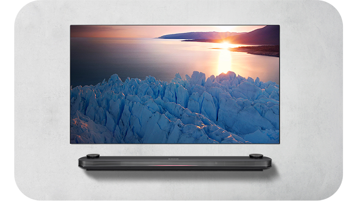벽에 설치된 LG TV 화면에 빙하와 해안선을 따라 해가 지는 장면이 선명하게 표시됨. TV 아래에는 얇은 사운드바가 벽면에 부착되어 있음.