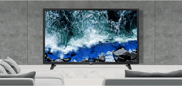 거실 공간에 배치된 LG TV가 테이블 위에 놓여 있으며, 화면에는 파도가 바위에 부딪히는 바다 풍경이 선명하게 표시됨. TV 앞에는 회색 톤의 소파와 쿠션이 배치되어 있고, 벽은 콘크리트 스타일의 모던한 디자인으로 구성됨.